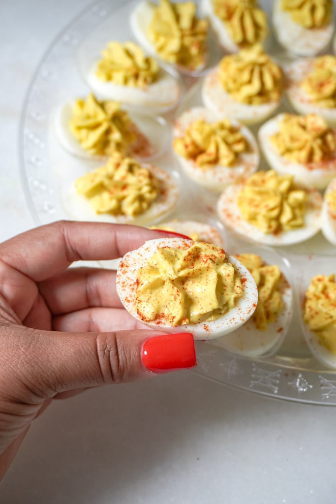a hand holding one deviled egg above a platter containing more