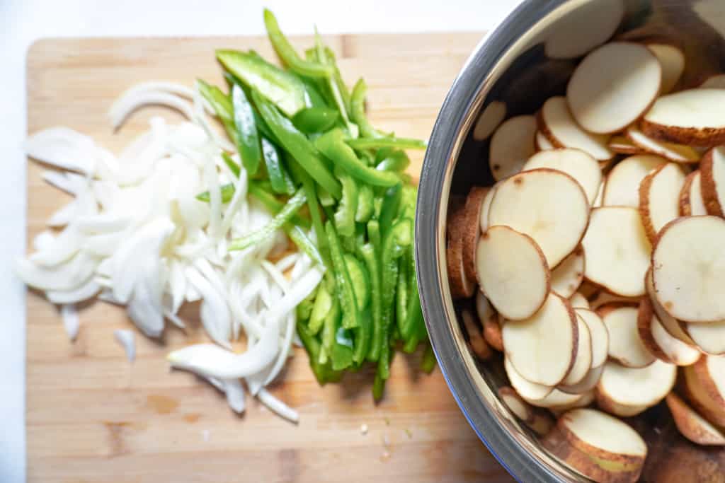 ingredients for smothered potatoes: sliced onions and peppers on a cutting board beside a bowl of sliced russet potatoes