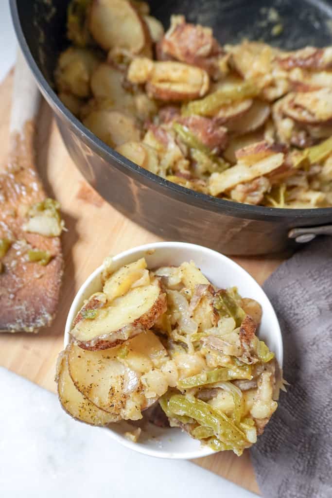 a small bowl full of smothered potatoes beside a pot containing more with a wooden spoon