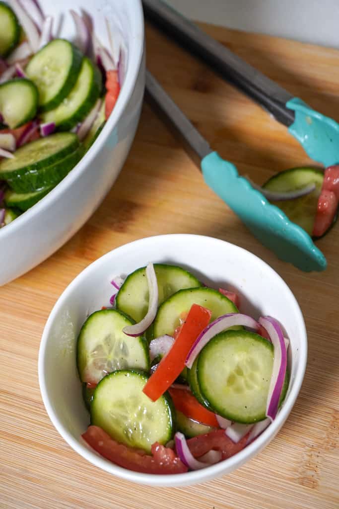 an individual serving of cucumber onion salad with a larger bowl in the background with teal tongs