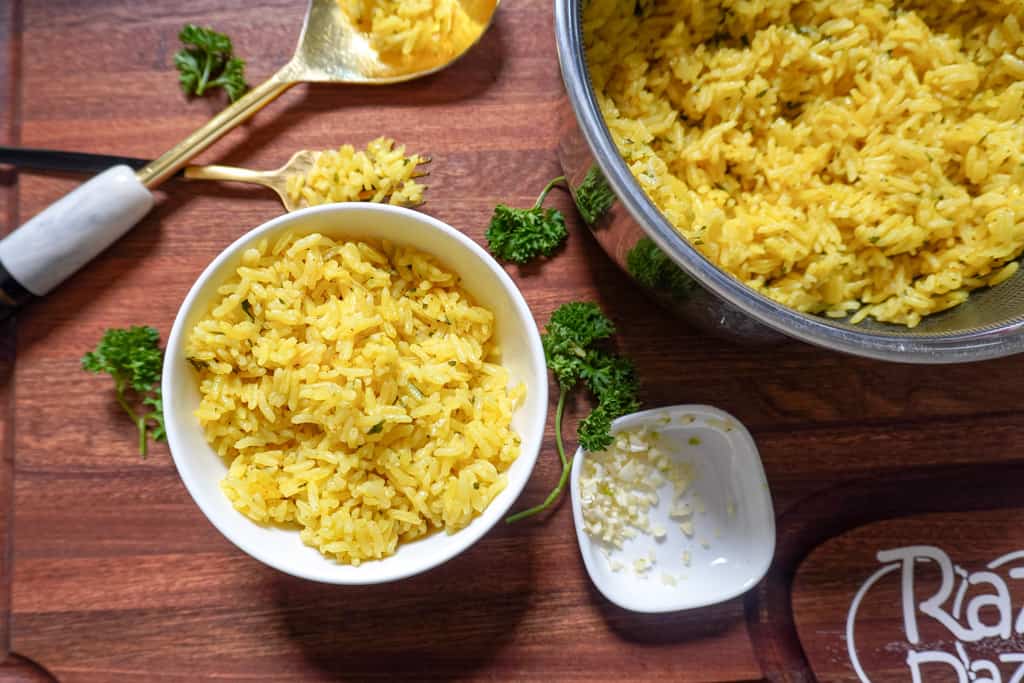 overhead view of a bowl of yellow greek rice next to a spoon, scattered herbs, and a pot containing more