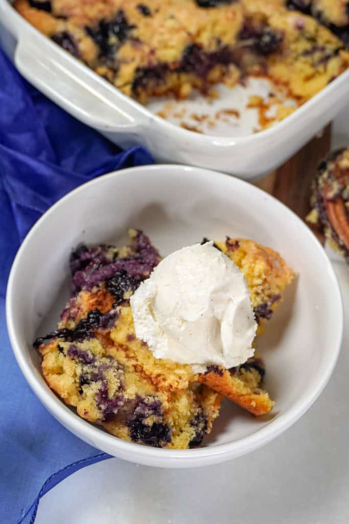 A bowl of blueberry cobbler topped with ice cream, next to the baking dish.