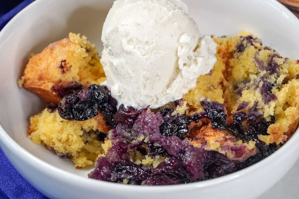 a close-up of a serving of blueberry cobbler in a white bowl, topped with a generous scoop of vanilla ice cream