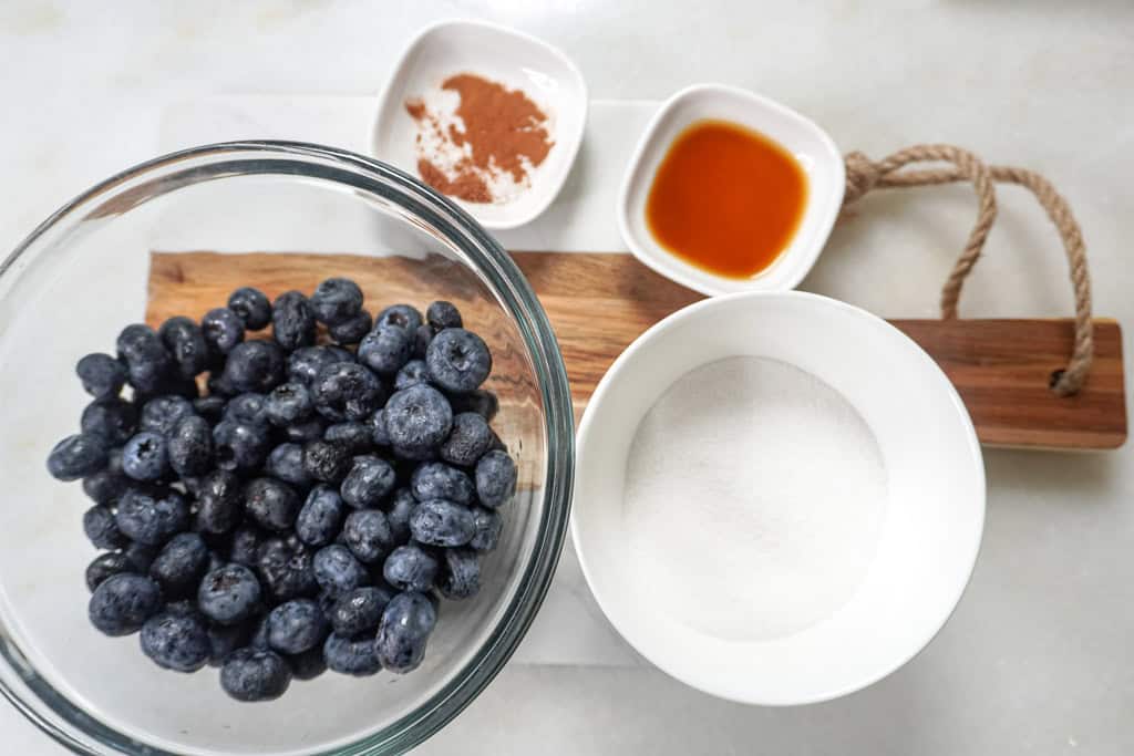 ingredients needed for the blueberry cobbler recipe: a bowl of blueberries, a stick of butter, and small saucers containing vanilla extract and cinnamon