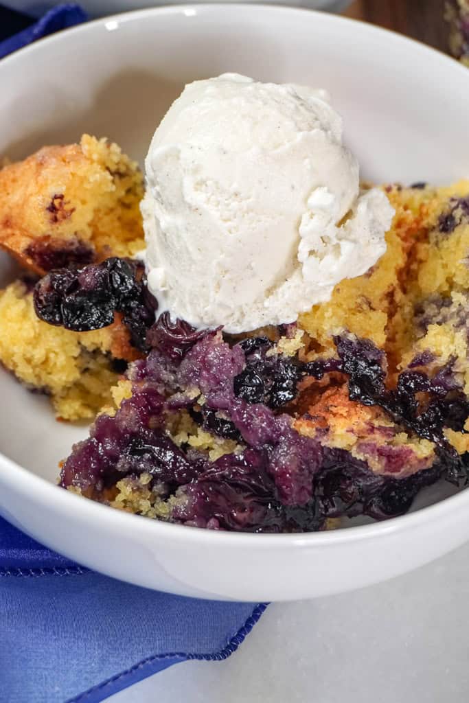 a close-up of a serving of blueberry cobbler in a white bowl, topped with a generous scoop of vanilla ice cream