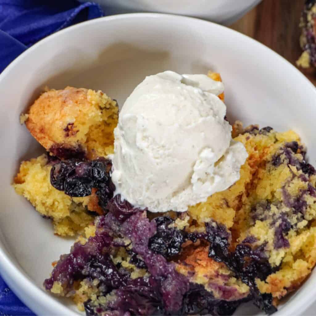 a serving of blueberry cobbler in a white bowl, topped with a scoop of vanilla ice cream