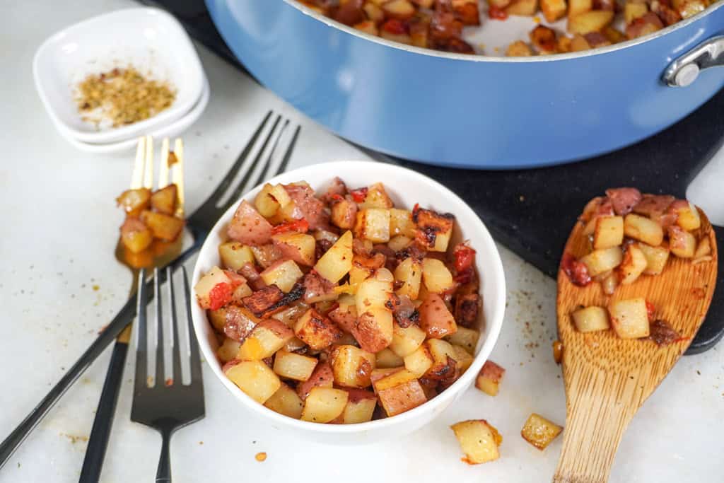 Another view of Potatoes O'Brien in a white bowl with a blue skillet and seasoning in the background.