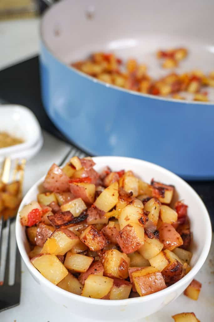 A white bowl filled with Potatoes O'Brien, placed near a blue skillet.