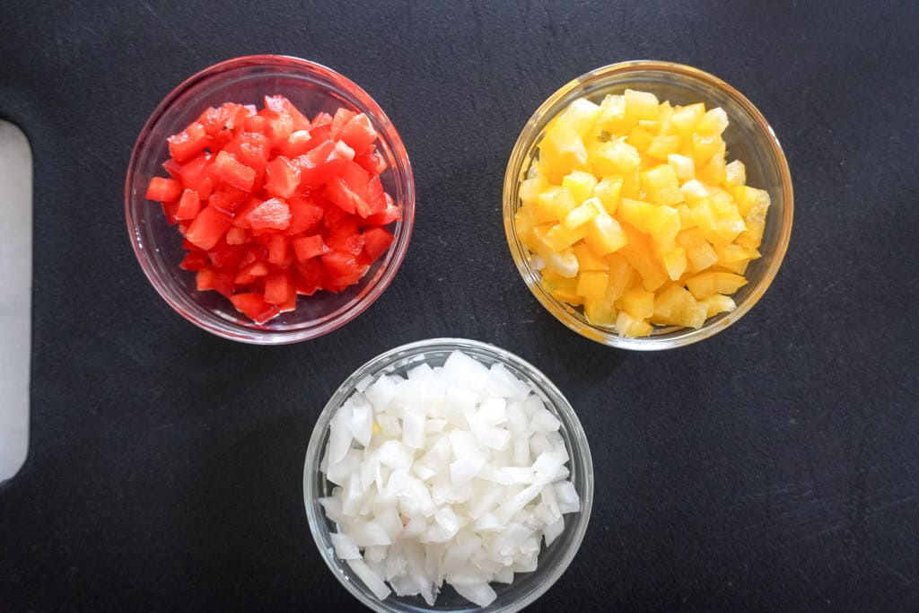 Diced red bell peppers, yellow bell peppers, and onions in separate glass bowls.