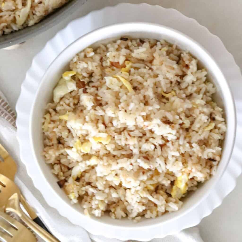 garlic fried rice with scrambled eggs in a white bowl beside a skillet and golden cutlery