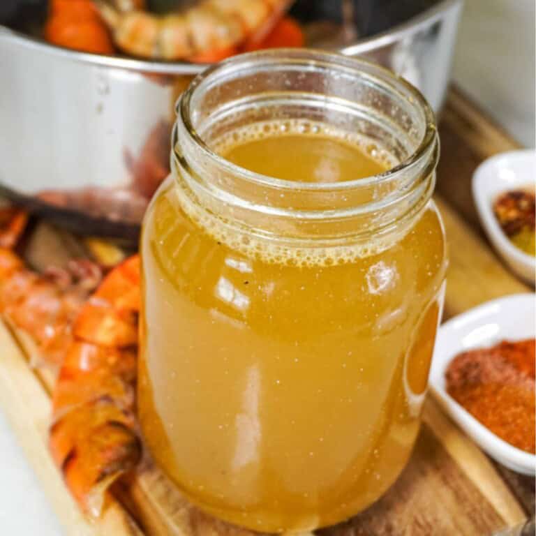 a jar of golden seafood stock sitting on a wooden surface, with shrimp shells and spices in the background, suggesting fresh ingredients used to make the stock