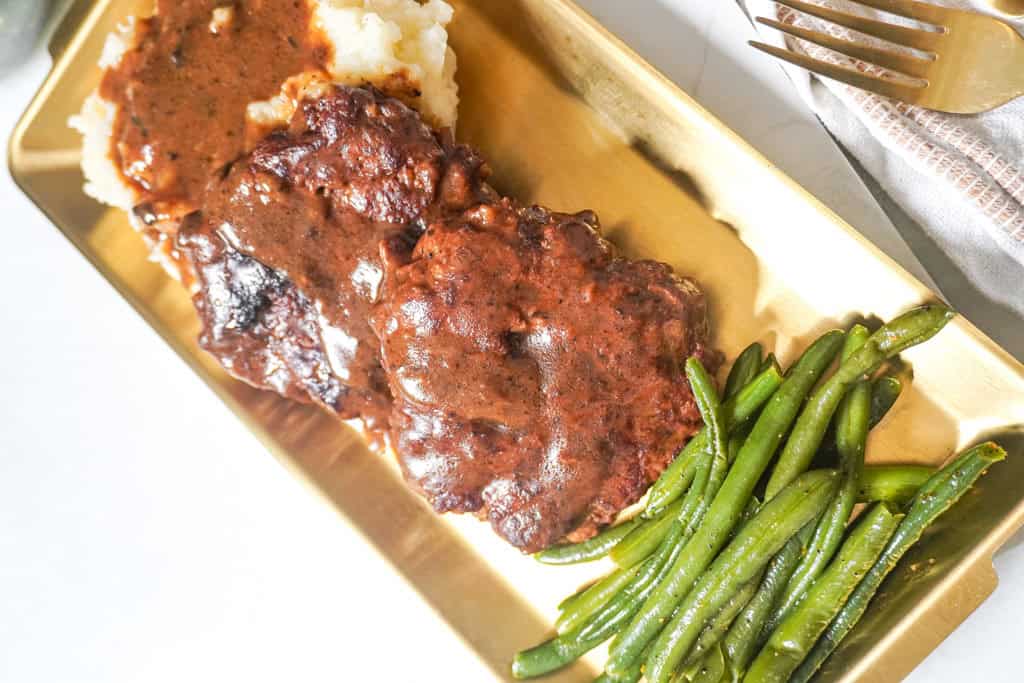 Side view of juicy hamburger steaks with brown gravy, mashed potatoes, and green beans on a gold platter.