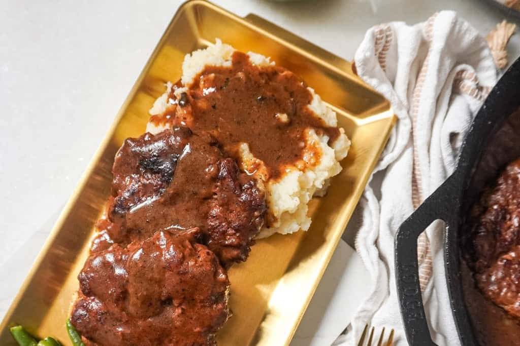 Mashed potatoes topped with brown gravy alongside hamburger steaks and green beans, served on a gold tray.