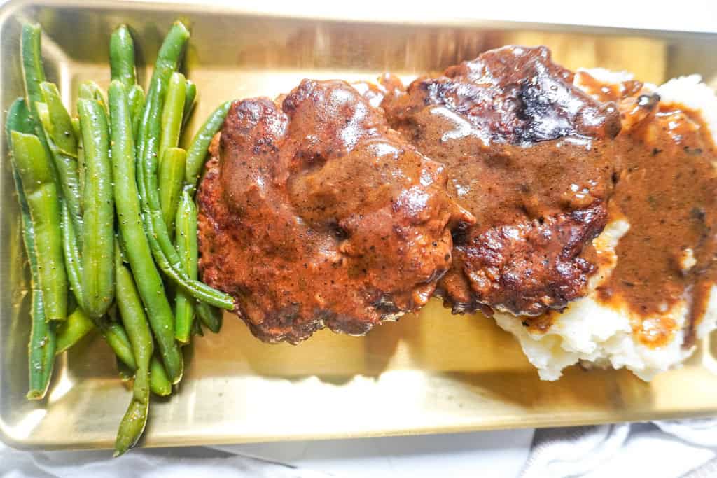Close-up of two gravy-smothered hamburger steaks with mashed potatoes and green beans, plated on a gold tray.