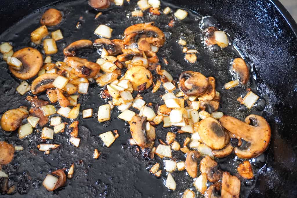 Chopped onions and sliced mushrooms sizzling in a black cast-iron skillet with oil.
