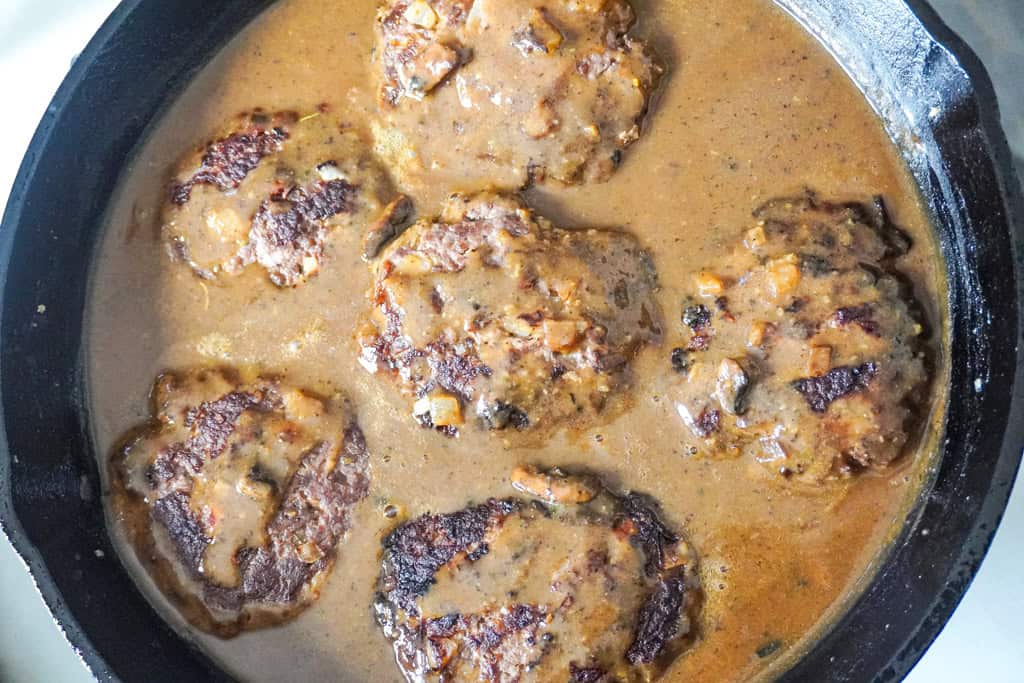 Golden-brown beef patties simmering in a rich brown gravy inside a cast-iron skillet.