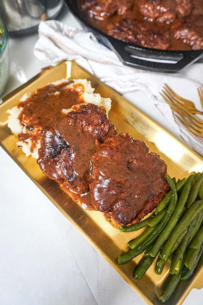 Overhead shot of a plate with two hamburger steaks smothered in brown gravy, mashed potatoes, and green beans, with a skillet of gravy in the background.