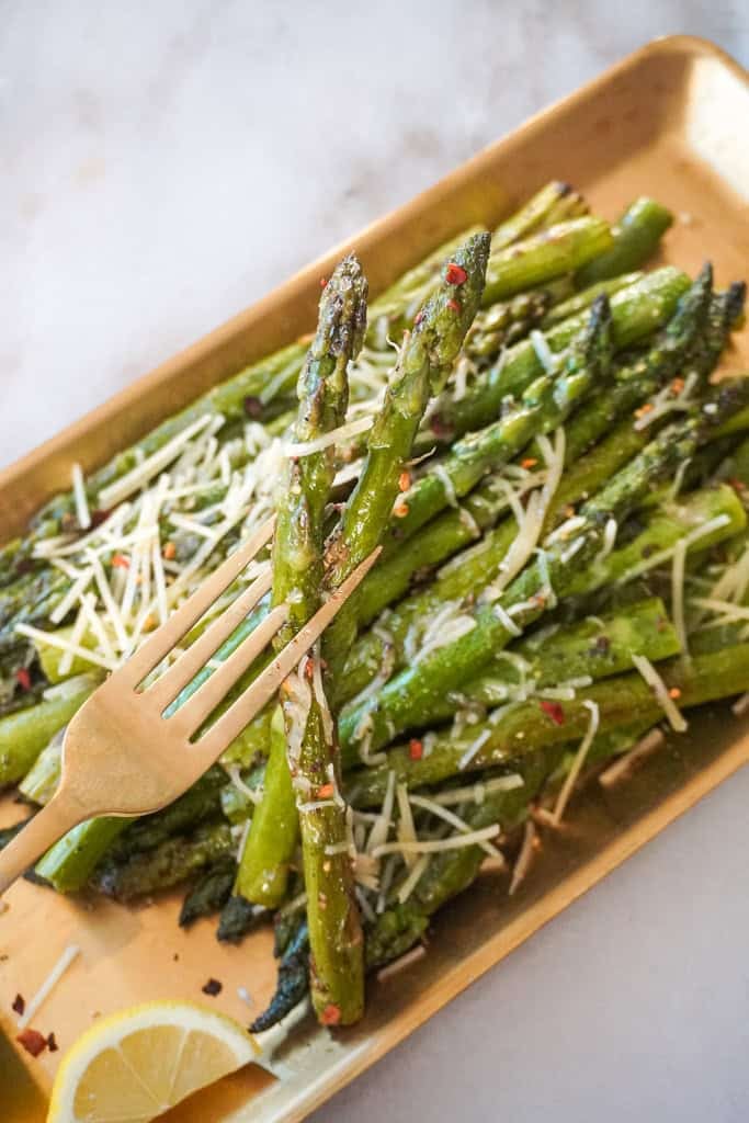 pan fried asparagus A close-up of a fork holding two spears of pan-fried asparagus with melted Parmesan and red pepper flakes, served on a gold tray.