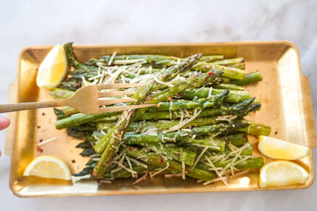 A close-up of a fork lifting a piece of garlic Parmesan asparagus from a gold tray, with crispy asparagus, melted cheese, and red pepper flakes in the background.
