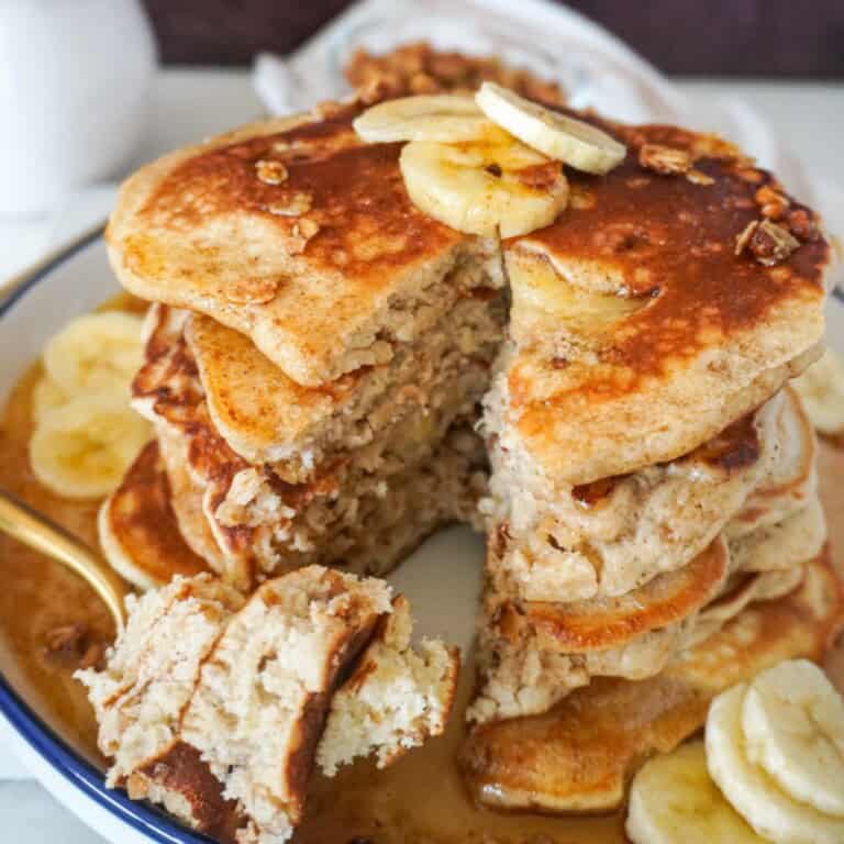 A stack of fluffy banana pancakes topped with banana slices and granola sits on a syrup-covered plate, with a bite cut out to show the soft, moist interior.